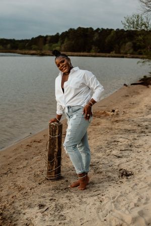 Figure in white shirt and jeans poses artistically on a sandy beach near water.