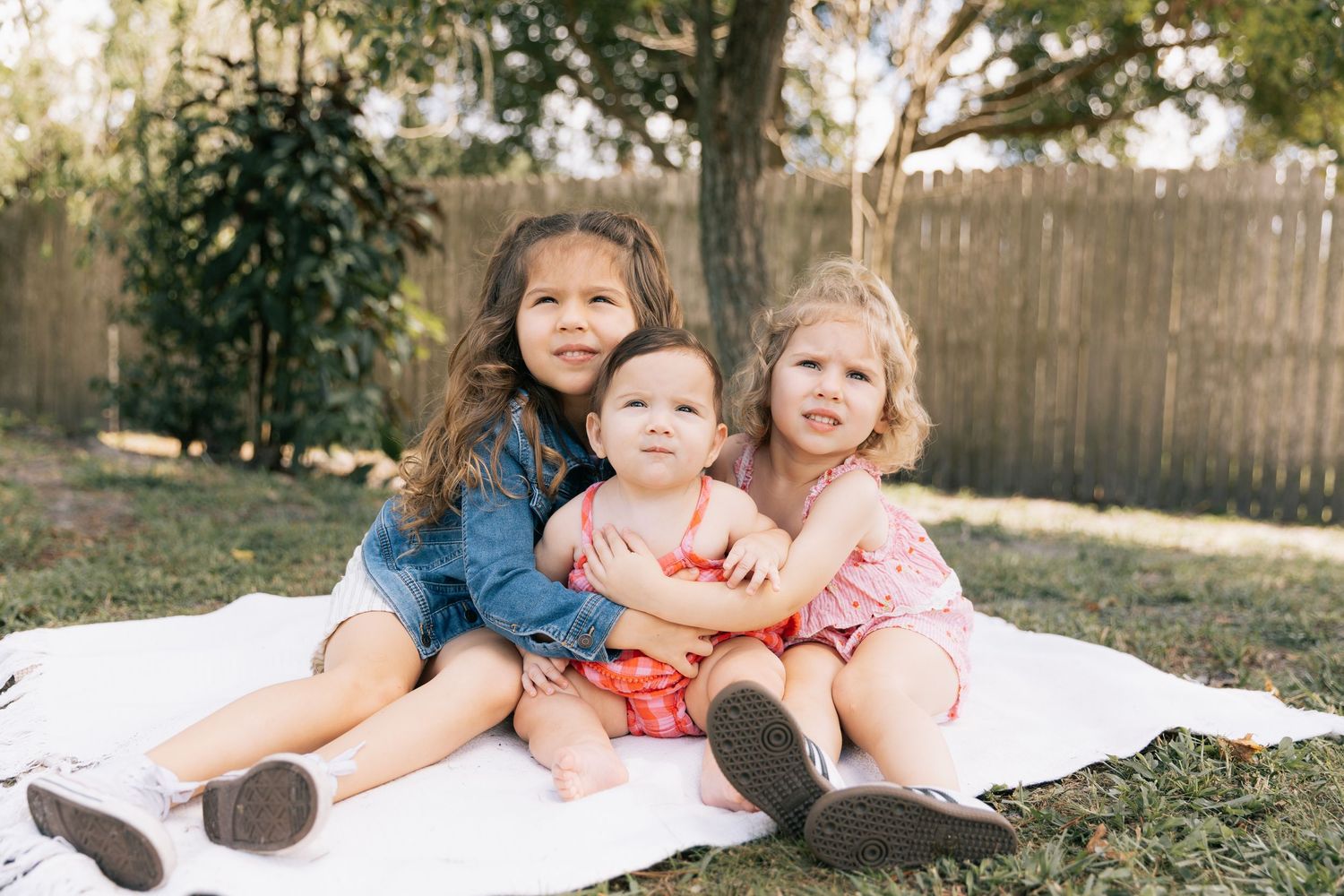 Three young girls cuddle on a white blanket in a sunny backyard with a wooden fence, including a baby in a red outfit.