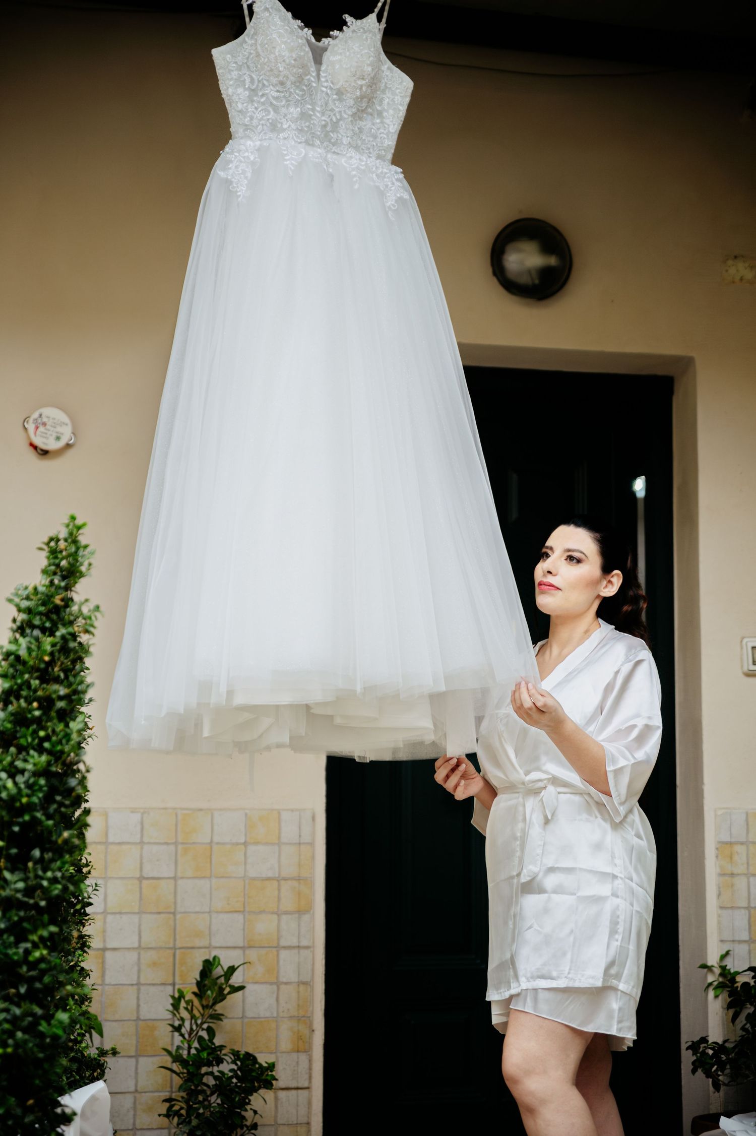 A beautiful white wedding dress hangs from above as someone in a silk robe admires it from below.