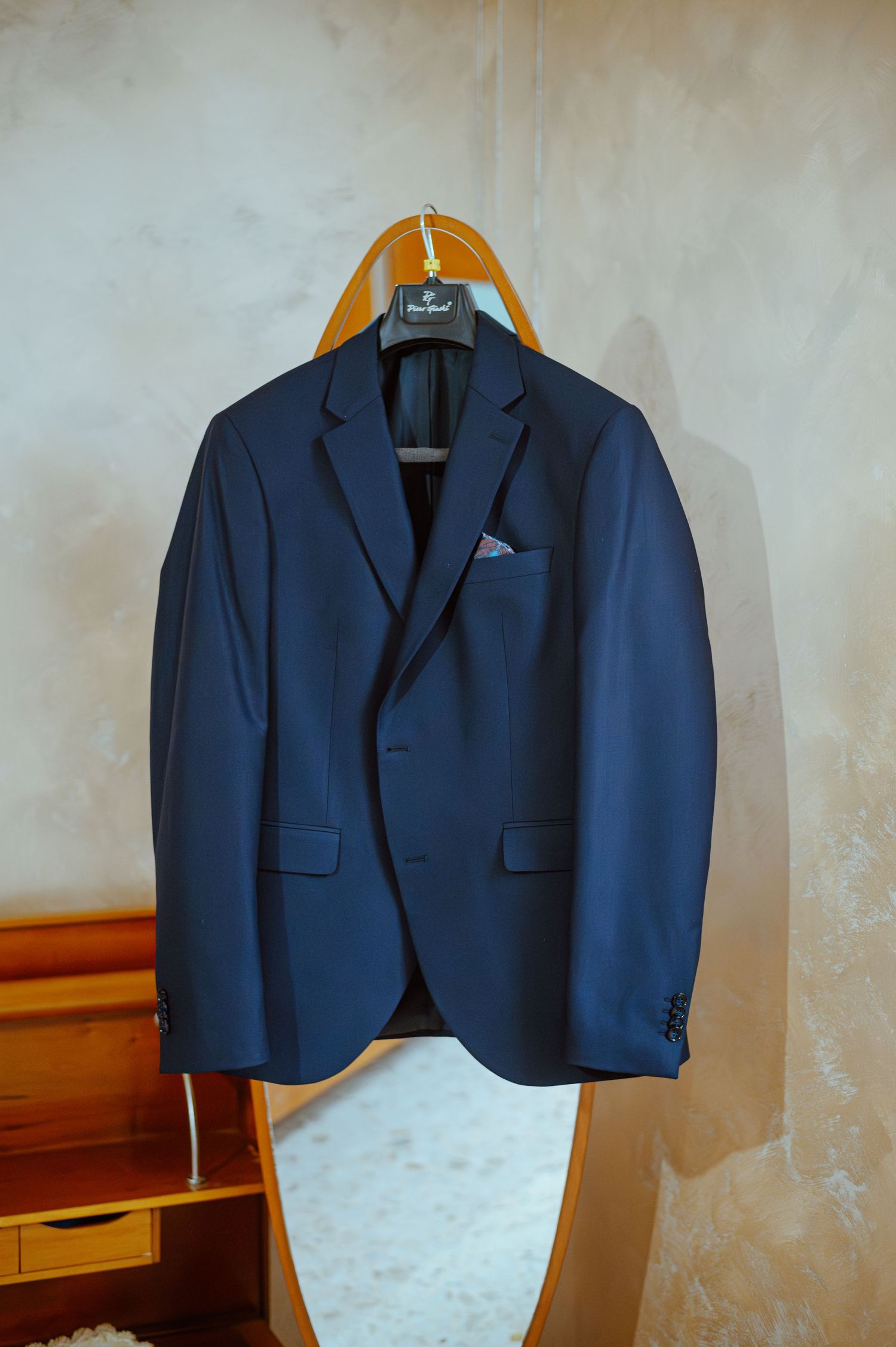 Navy blue formal suit jacket hanging on wooden hanger against beige wall.