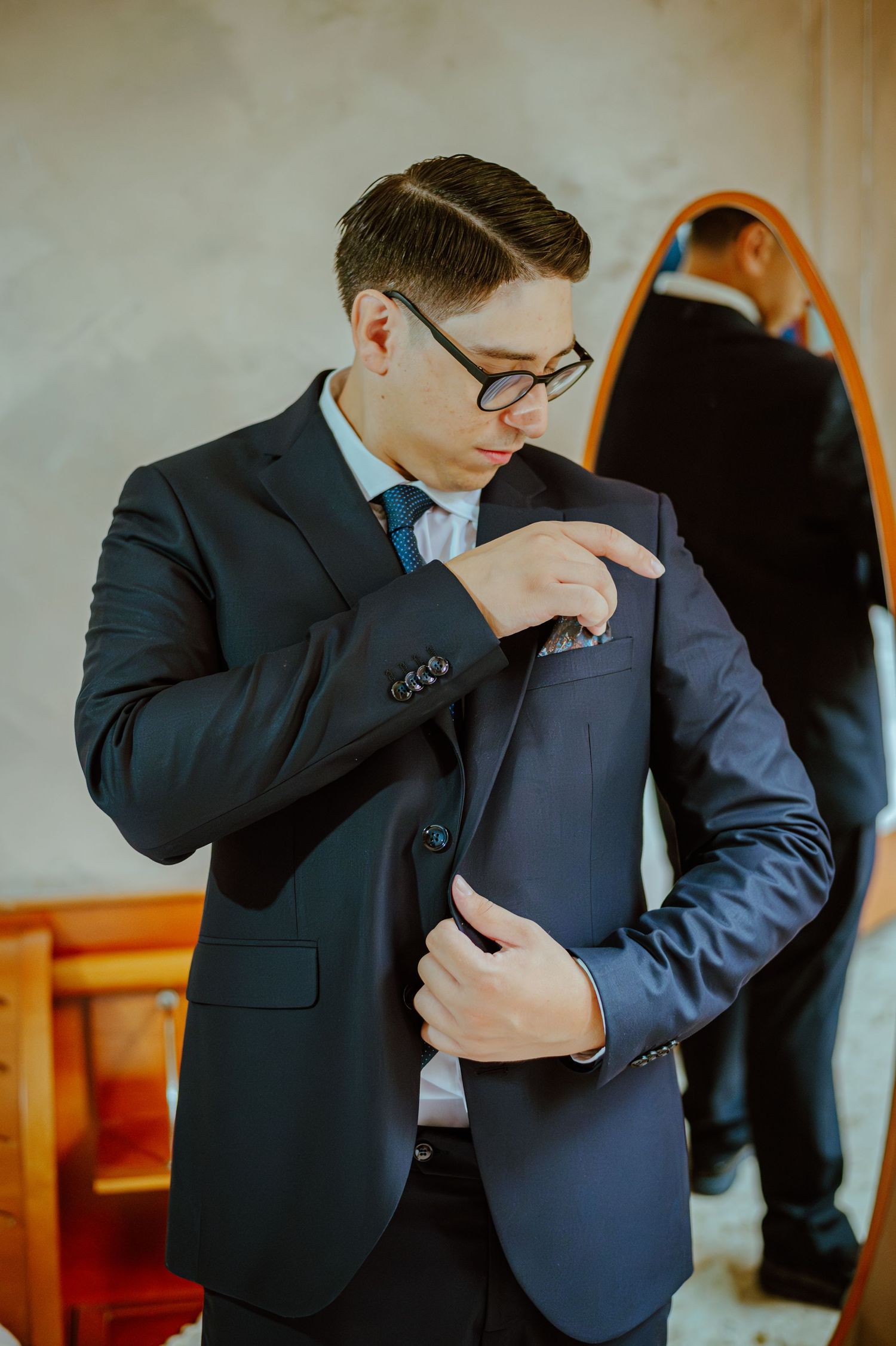 A person in a dark suit and glasses adjusting formal attire in front of a mirror.