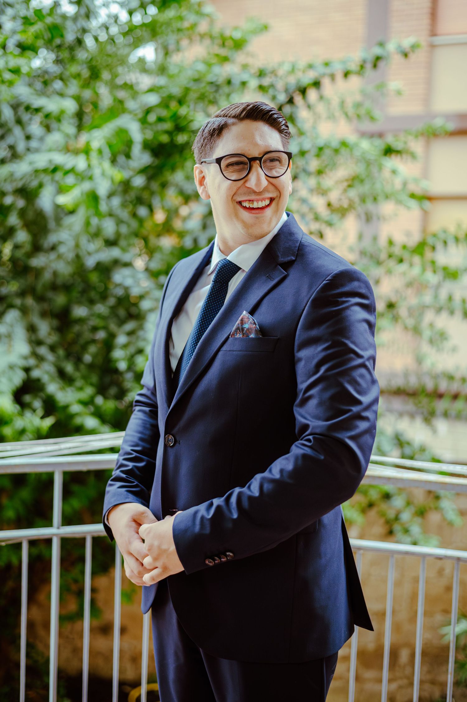 A person in navy suit and glasses standing on outdoor balcony during formal event.
