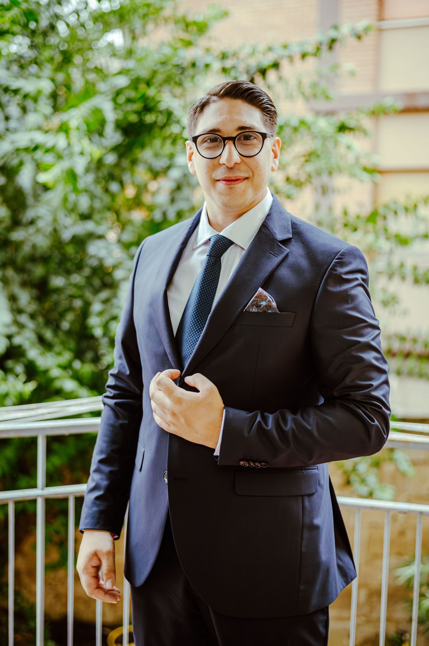 A person in fitted navy suit with pocket square posing on balcony with greenery backdrop.
