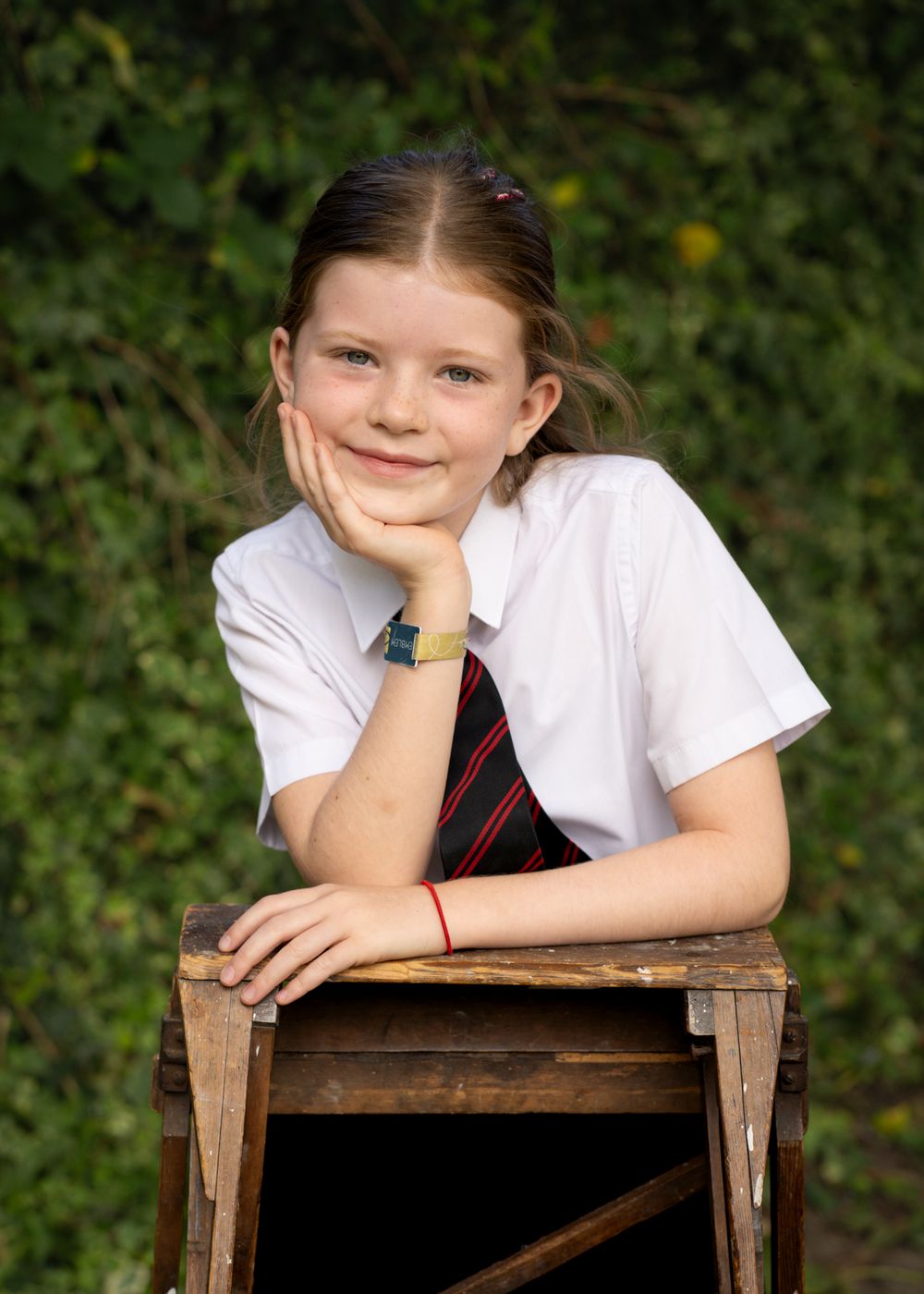 A child in a white shirt and red tie poses playfully on a wooden crate against a green leafy background.