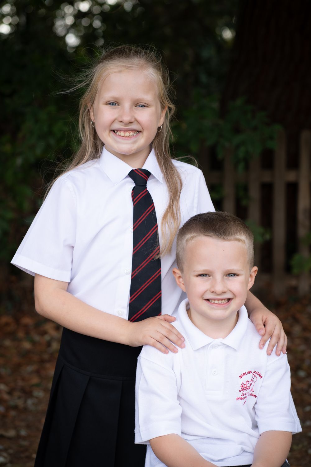 Two children in white school uniforms with striped ties pose together outdoors against a dark background.