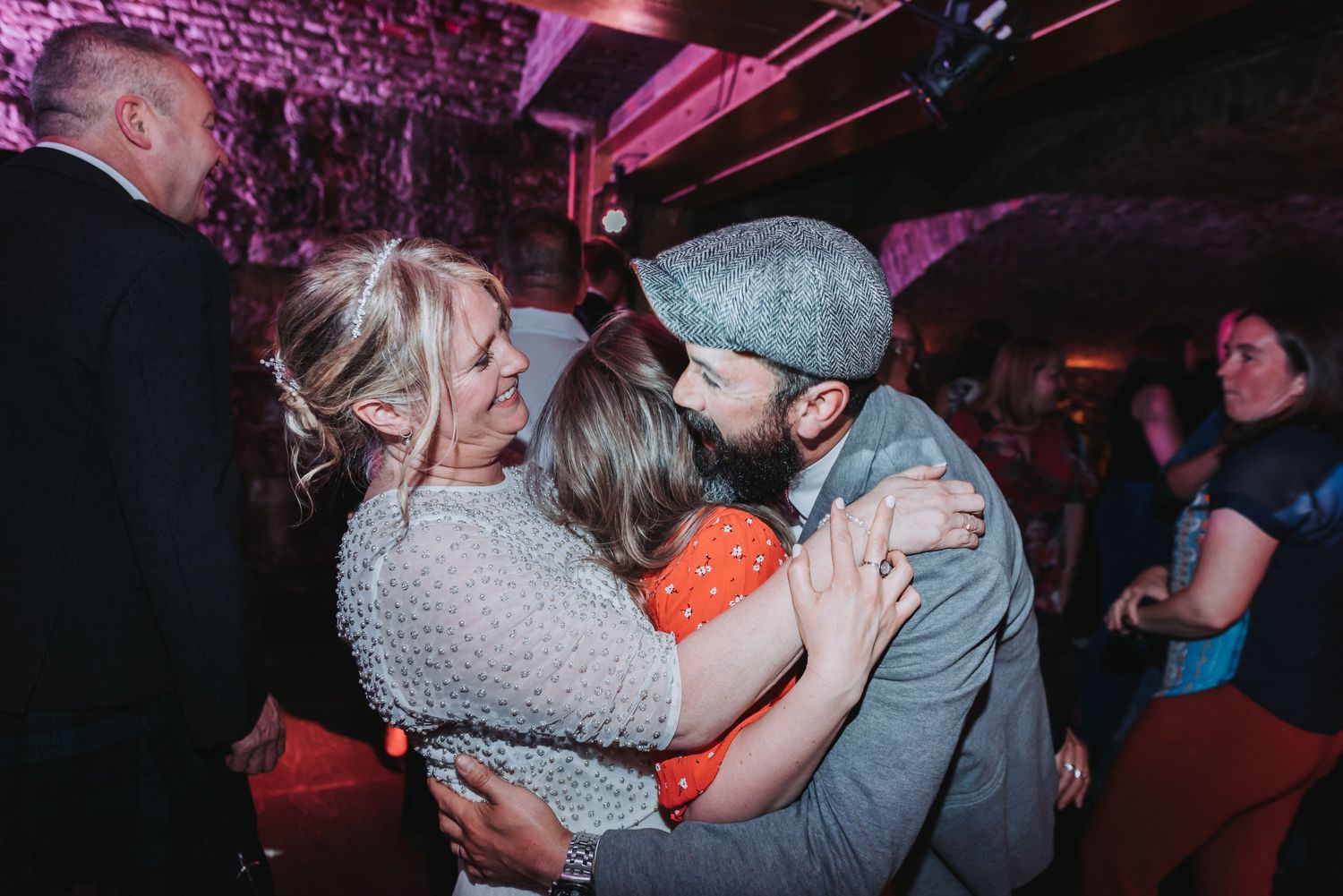 Couples dancing closely in a venue with pink-purple lighting and stone walls.
