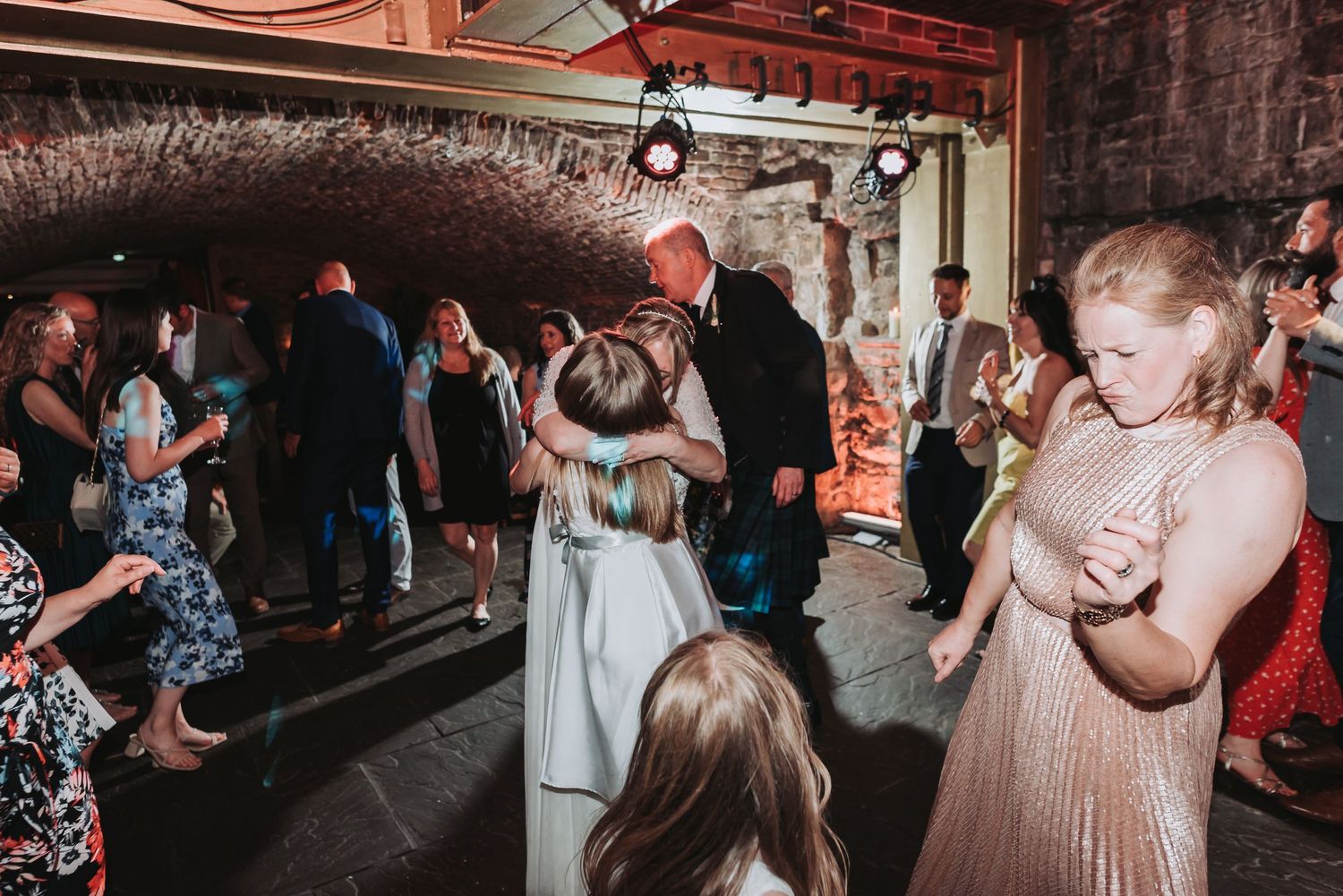 Wedding guests dancing in a rustic venue with exposed stone walls and overhead lighting.