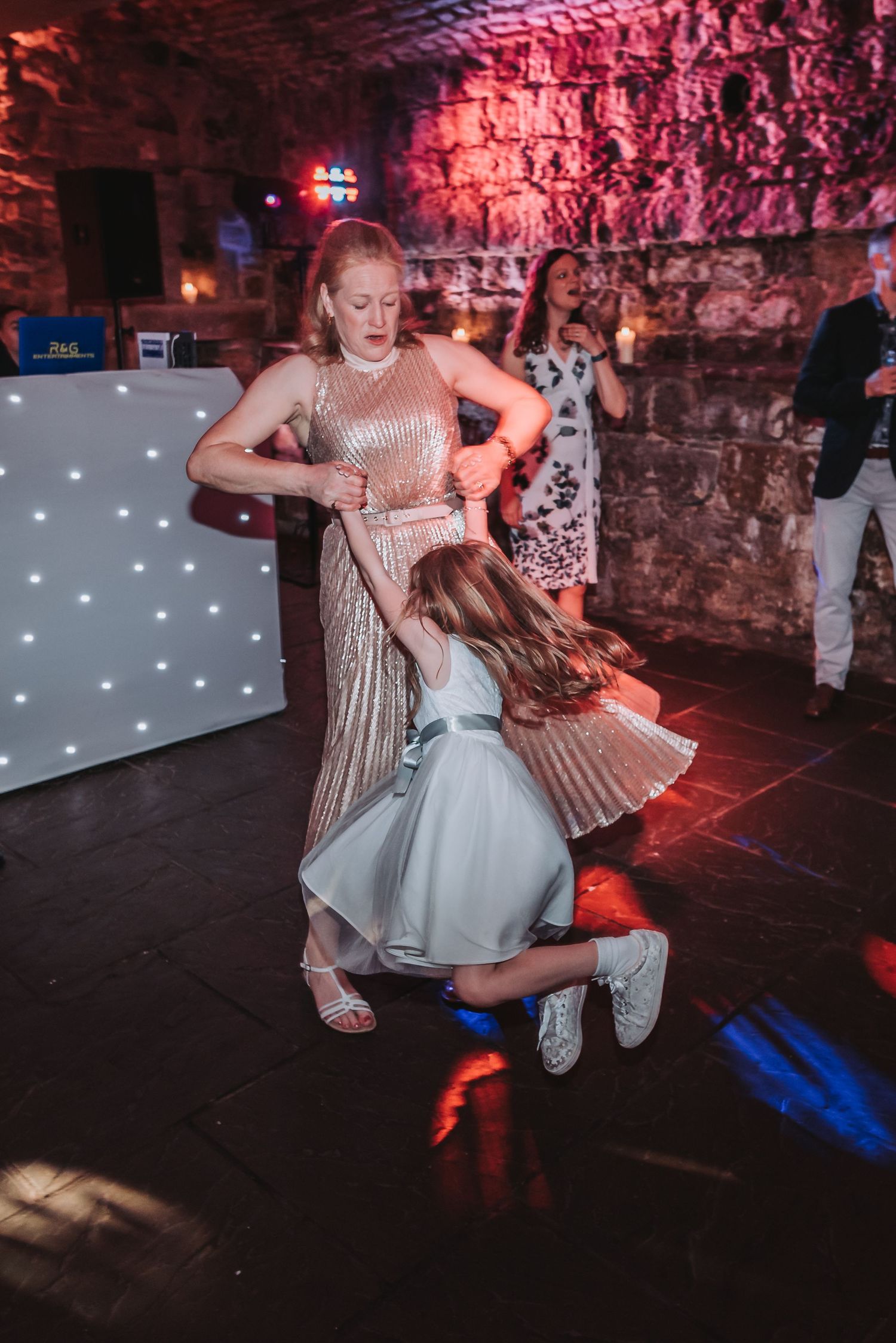 Dancing scene at a celebration with decorative lighting and stone wall backdrop.
