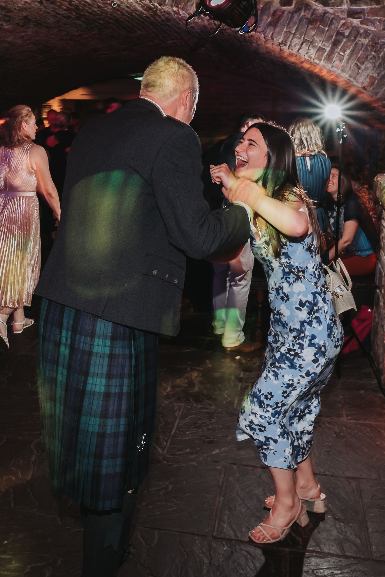 Guests dancing energetically at a wedding reception in a vaulted cellar with colorful lighting and traditional Scottish attire.