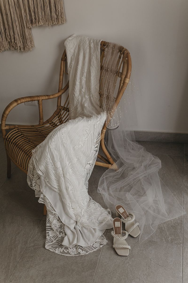 Lace wedding dress draped over rattan chair with bridal heels on tile floor, boho wedding styling.