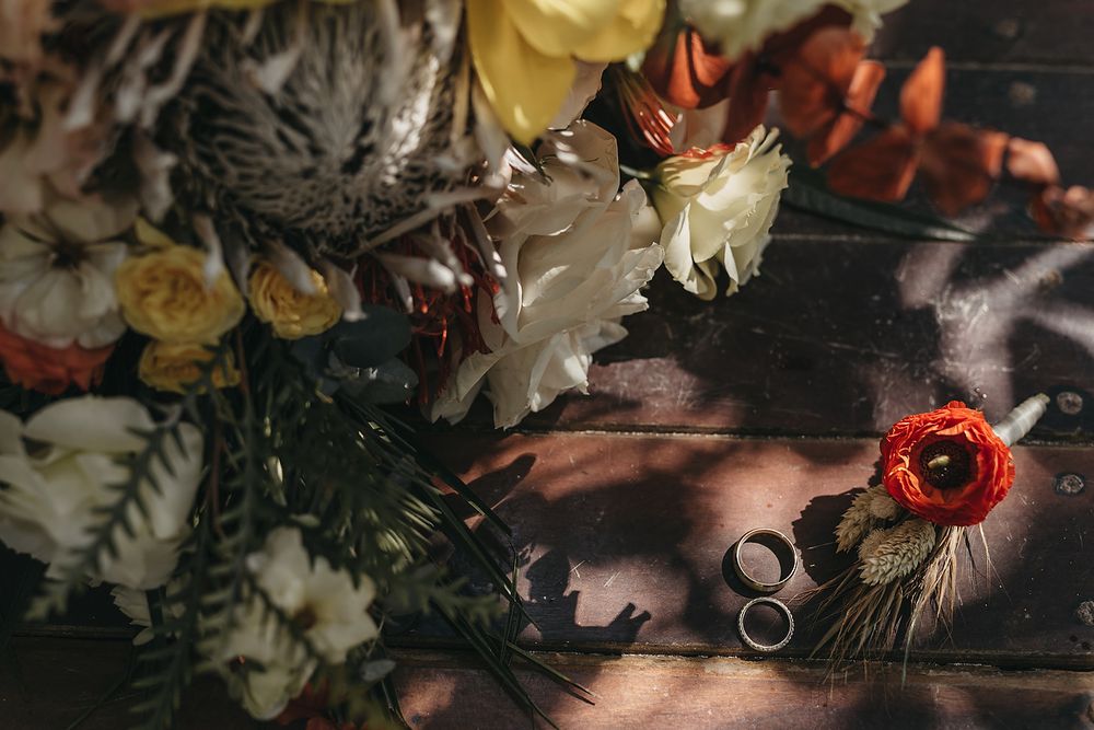 Wedding rings and floral bouquet with orange, white, and yellow flowers on a dark wooden surface.