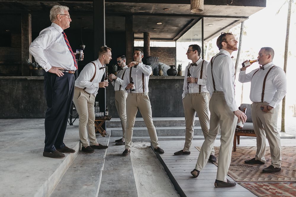 Groomsmen in suspenders and dress shirts socializing before a wedding ceremony in a rustic venue.