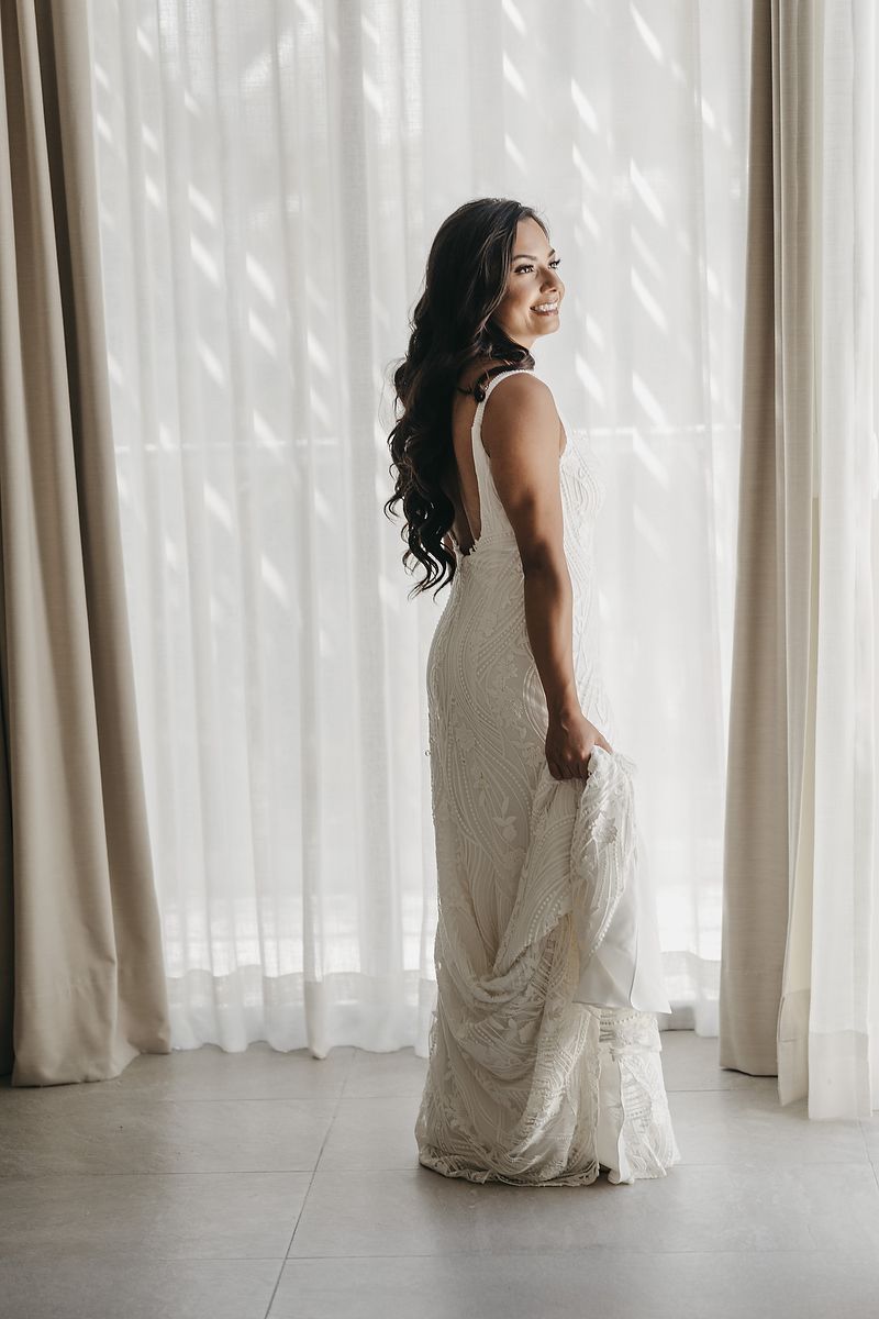 Bride in white beaded gown holding dress train, smiling over shoulder near sheer curtains in elegant bridal portrait.