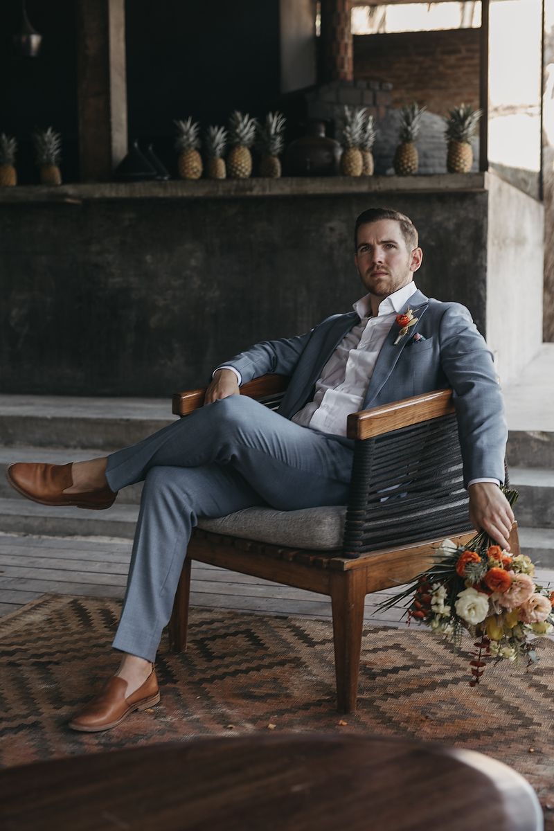 A groom in a light blue suit with a floral boutonniere sits relaxed in a wooden chair beside a bouquet.