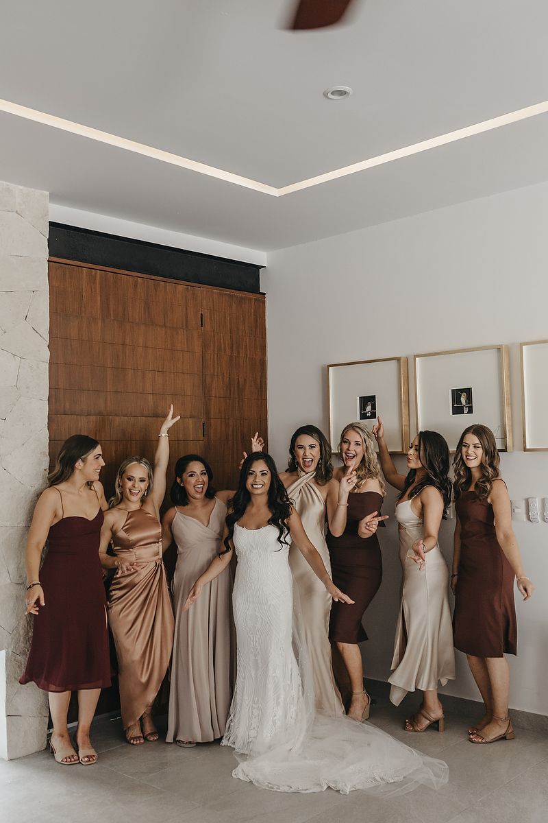 Bride in white lace gown poses with bridesmaids in mismatched earth-tone dresses in elegant modern interior.