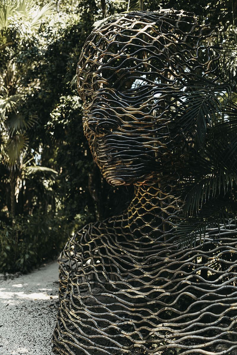 Wire mesh sculpture of a human figure surrounded by lush tropical greenery in an outdoor garden setting.