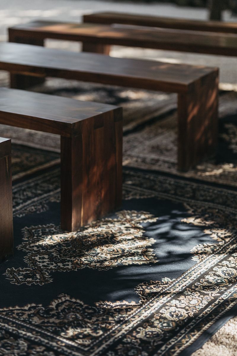 Dark wooden benches arranged on ornate patterned rugs outdoors, casting shadows in warm sunlight at a rustic wedding ceremony.