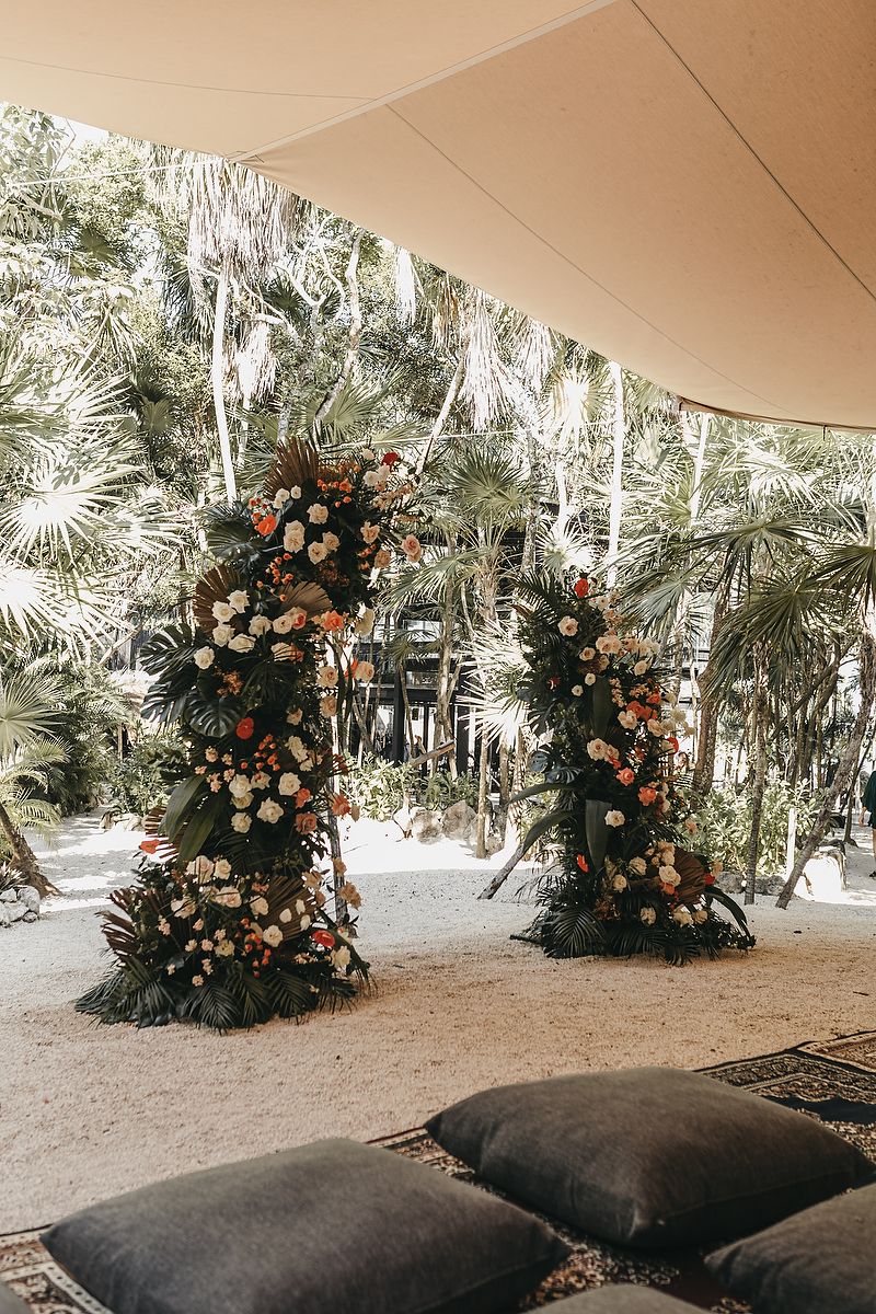 Tropical wedding ceremony arch with white and orange flowers, lush palm trees, and dark floor cushions on sandy ground.