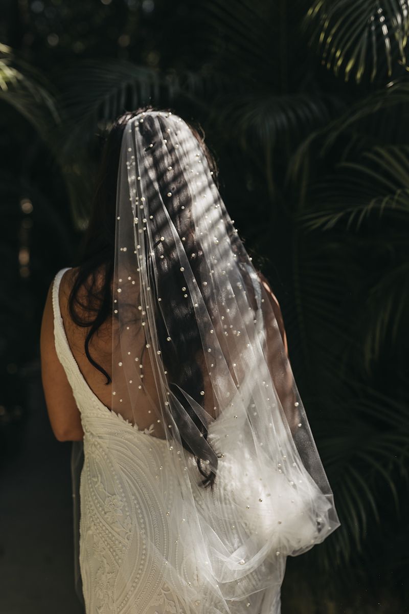 Bride wearing a pearl-embellished wedding veil and lace gown, photographed from behind against tropical palm leaves.