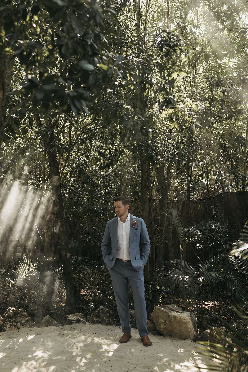 A groom in a blue suit with a boutonniere stands confidently in a lush tropical forest setting with sunlight filtering through trees.