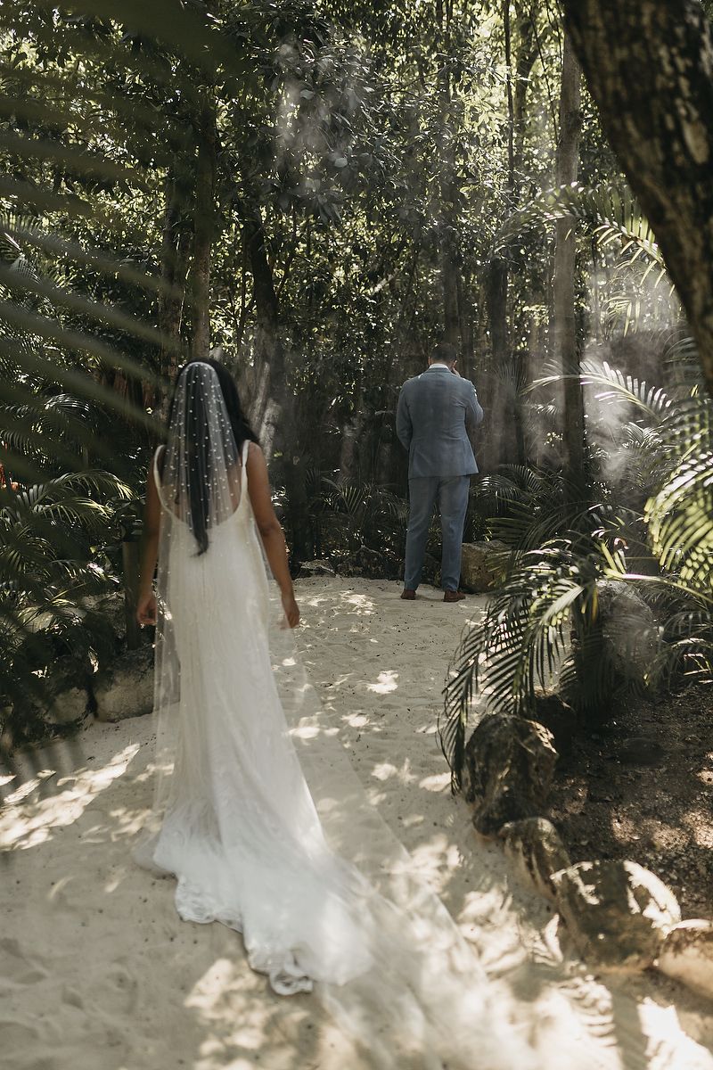 Bride in white gown and veil approaches groom in lush tropical garden for romantic first look wedding moment.