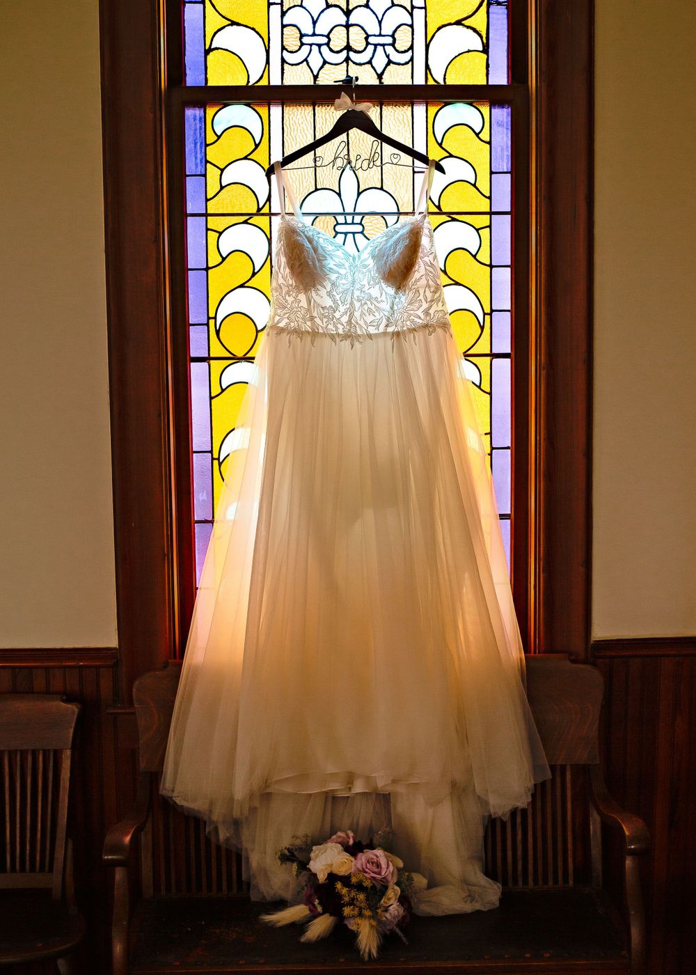 Beautiful white wedding dress hanging in front of a stained glass window with a bouquet at the base.