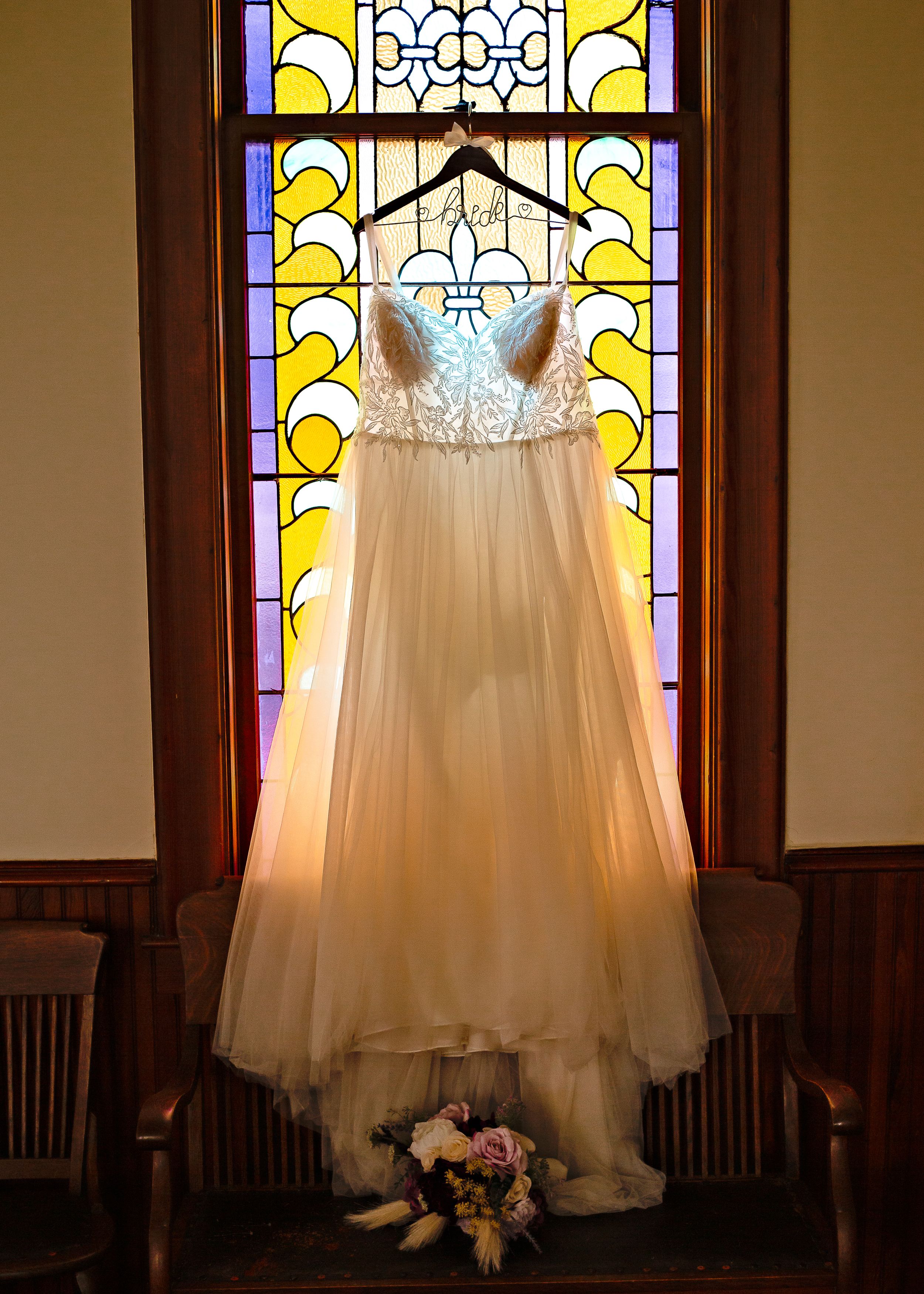 Beautiful white wedding dress hanging in front of a stained glass window with a bouquet at the base.
