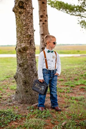 Young ring bearer in suspenders and bowtie poses confidently by a tree trunk.
