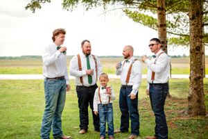 Group of groomsmen in casual attire with suspenders gathered under a tree.