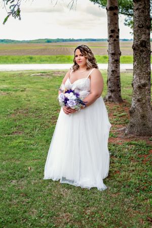 Bride in flowing white dress holds purple bouquet while standing under trees.