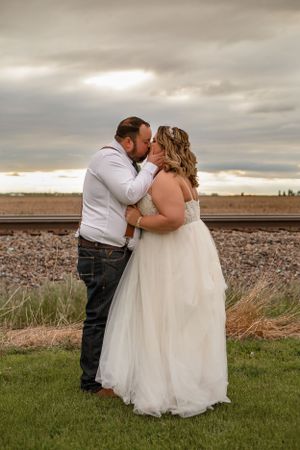 A tender moment between newlyweds against a dramatic cloudy sky by railroad tracks.