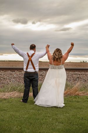 Joyful newlyweds celebrate with raised arms near railroad tracks under cloudy skies.