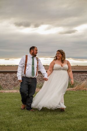 Happy couple walking and laughing together on grass near railroad tracks under dramatic skies.