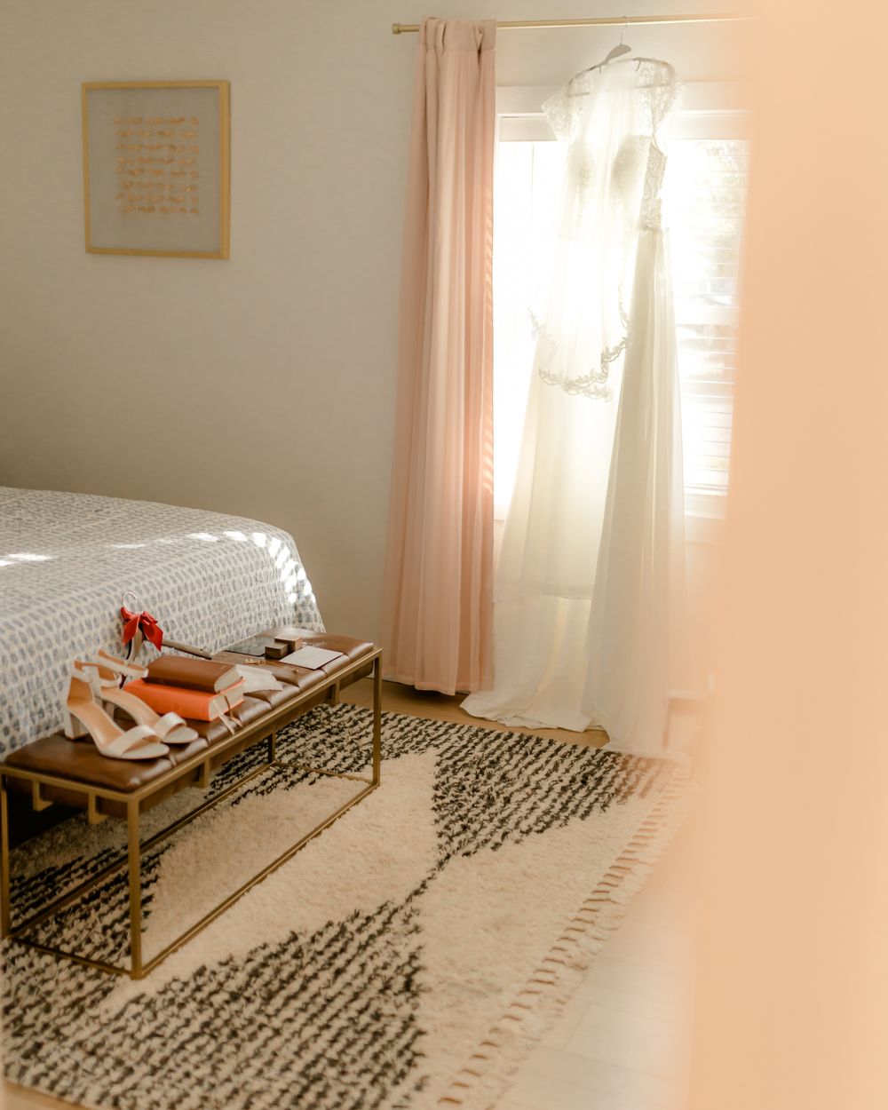 Sunlit bedroom with wedding dress hanging by window and accessories on a bench.