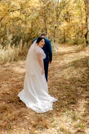 A wedding couple walking on a sunlit autumn forest path with the bride's flowing white dress trailing behind.