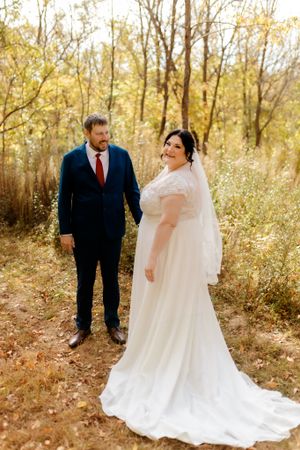 Elegant wedding portrait on a leaf-covered path with autumn trees in background.