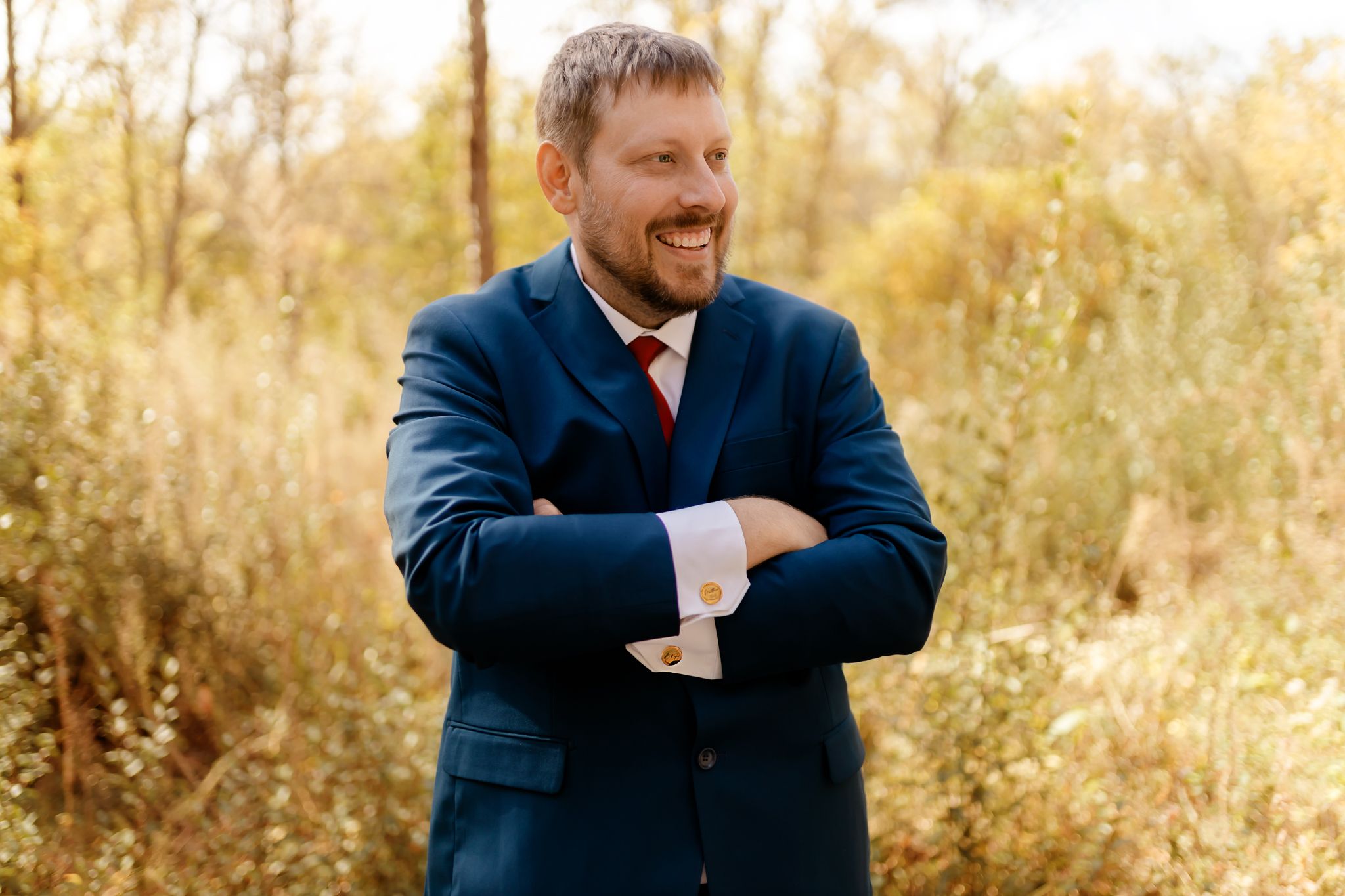 A groom in a navy suit smiles warmly while standing in a golden autumn field.
