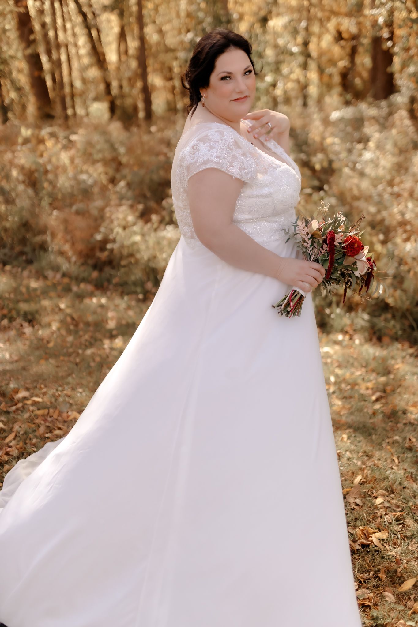 A bride in a lace-detailed wedding gown holds a red and white bouquet in an autumn forest setting.