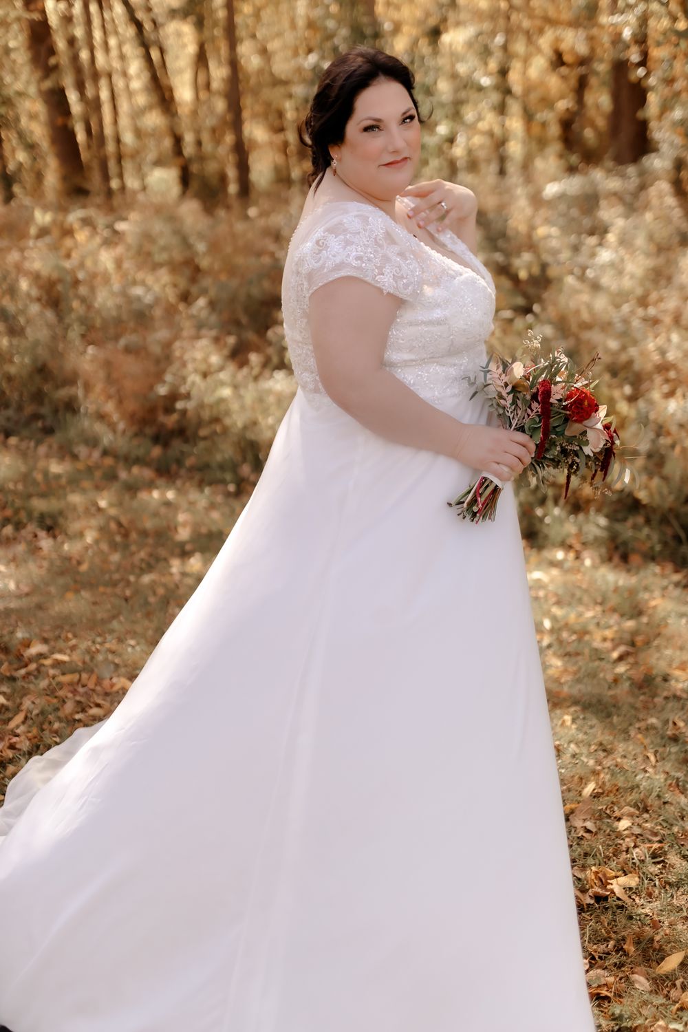 A bride in a lace-detailed wedding gown holds a red and white bouquet in an autumn forest setting.