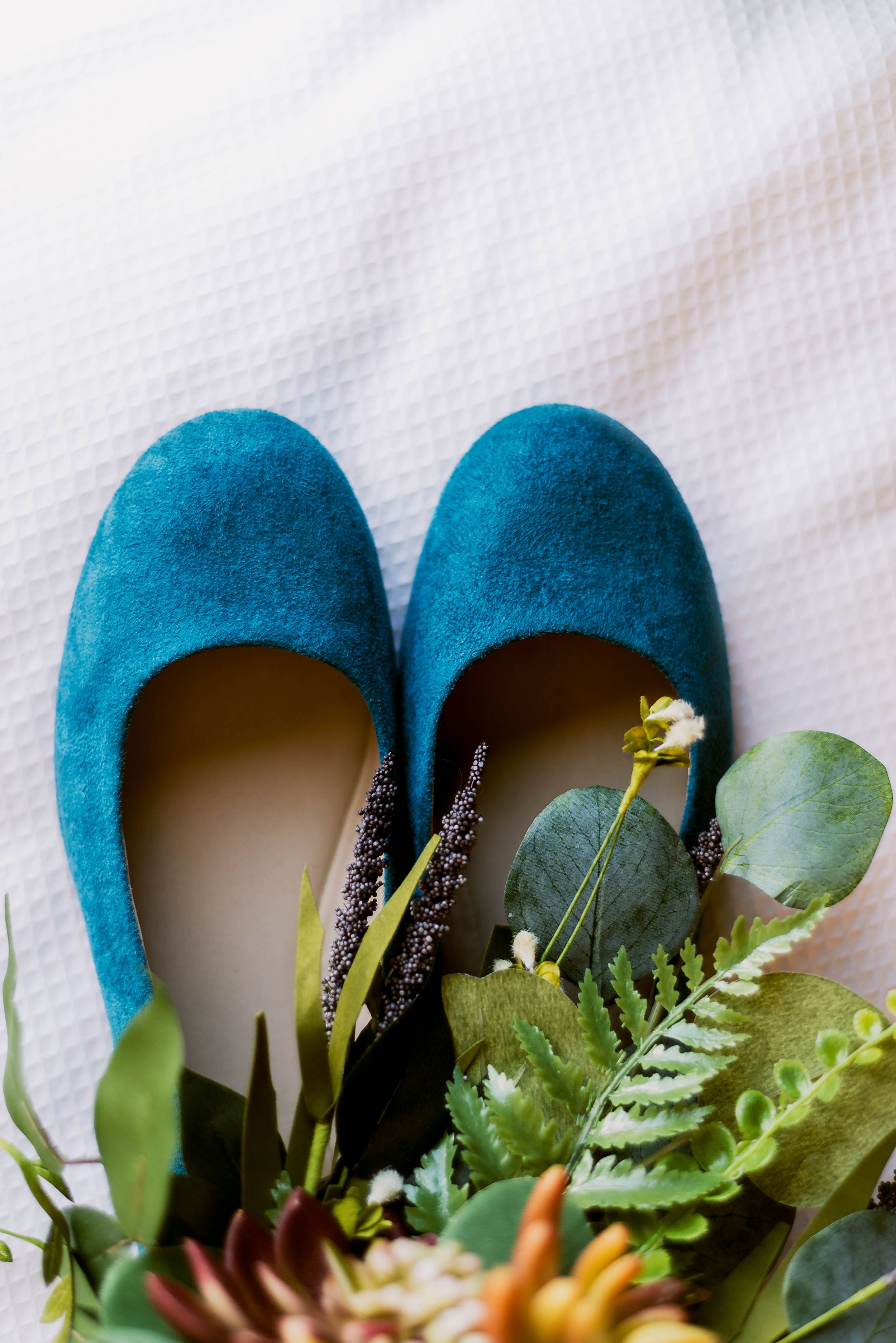 Teal blue suede flats with greenery and floral decorations on white background.