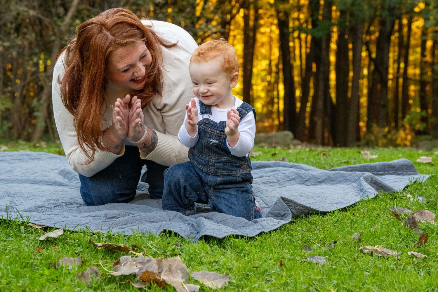 A parent and child share a tender moment while lying on a blanket outdoors during autumn.