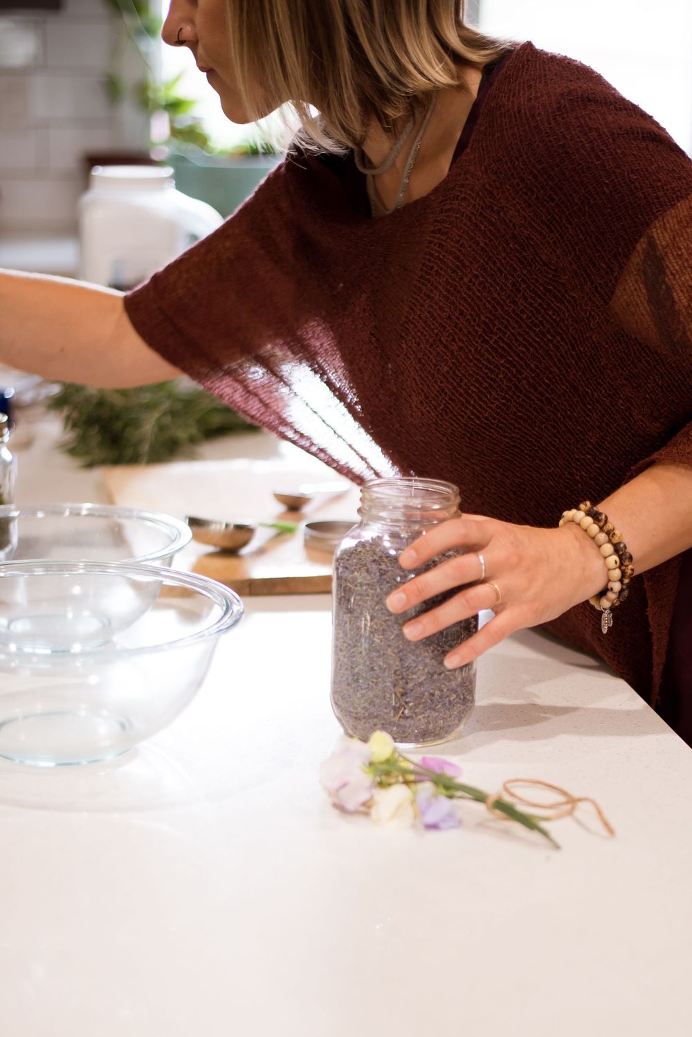 Someone in a burgundy sweater pouring ingredients into a glass jar on a white counter.
