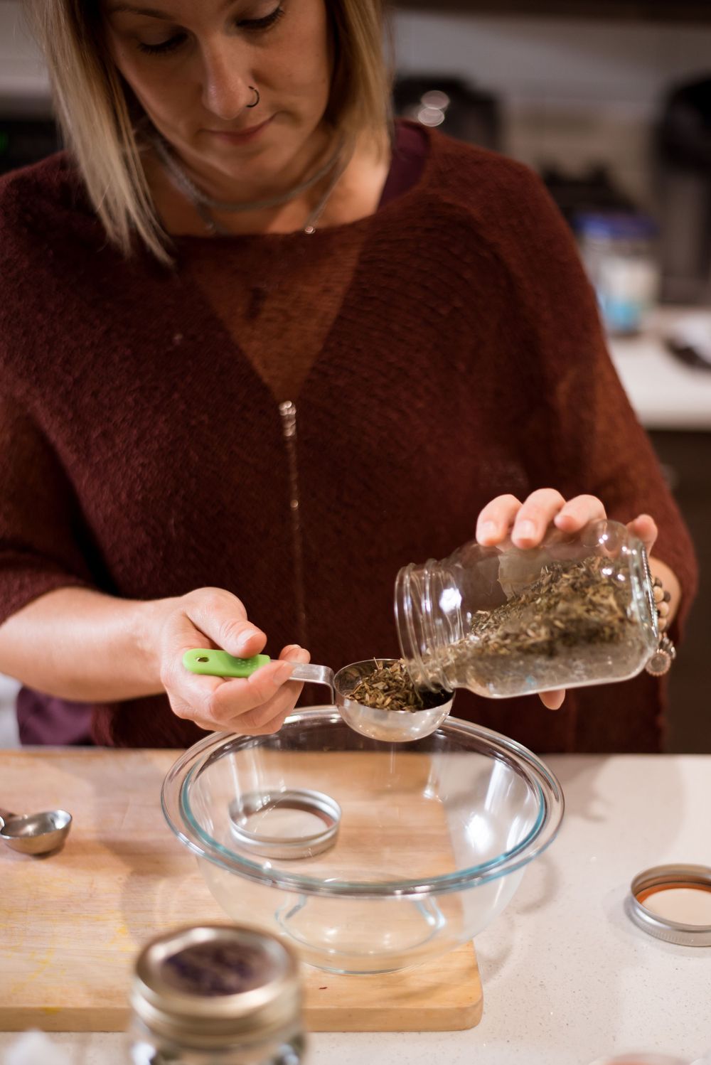 Measuring dried herbs from a mason jar into a glass bowl on a wooden cutting board.