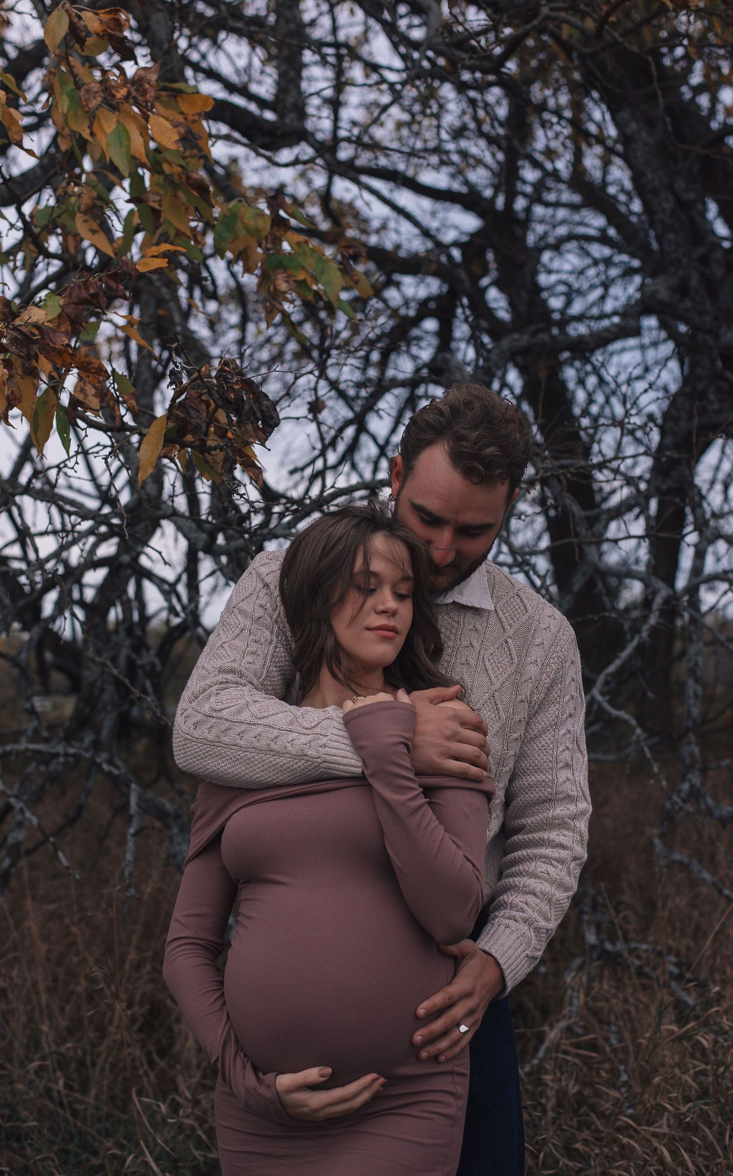 A dreamy, dark scene with a man holding a pregnant woman from behind, while she holds his arm and her belly. Couple Photography