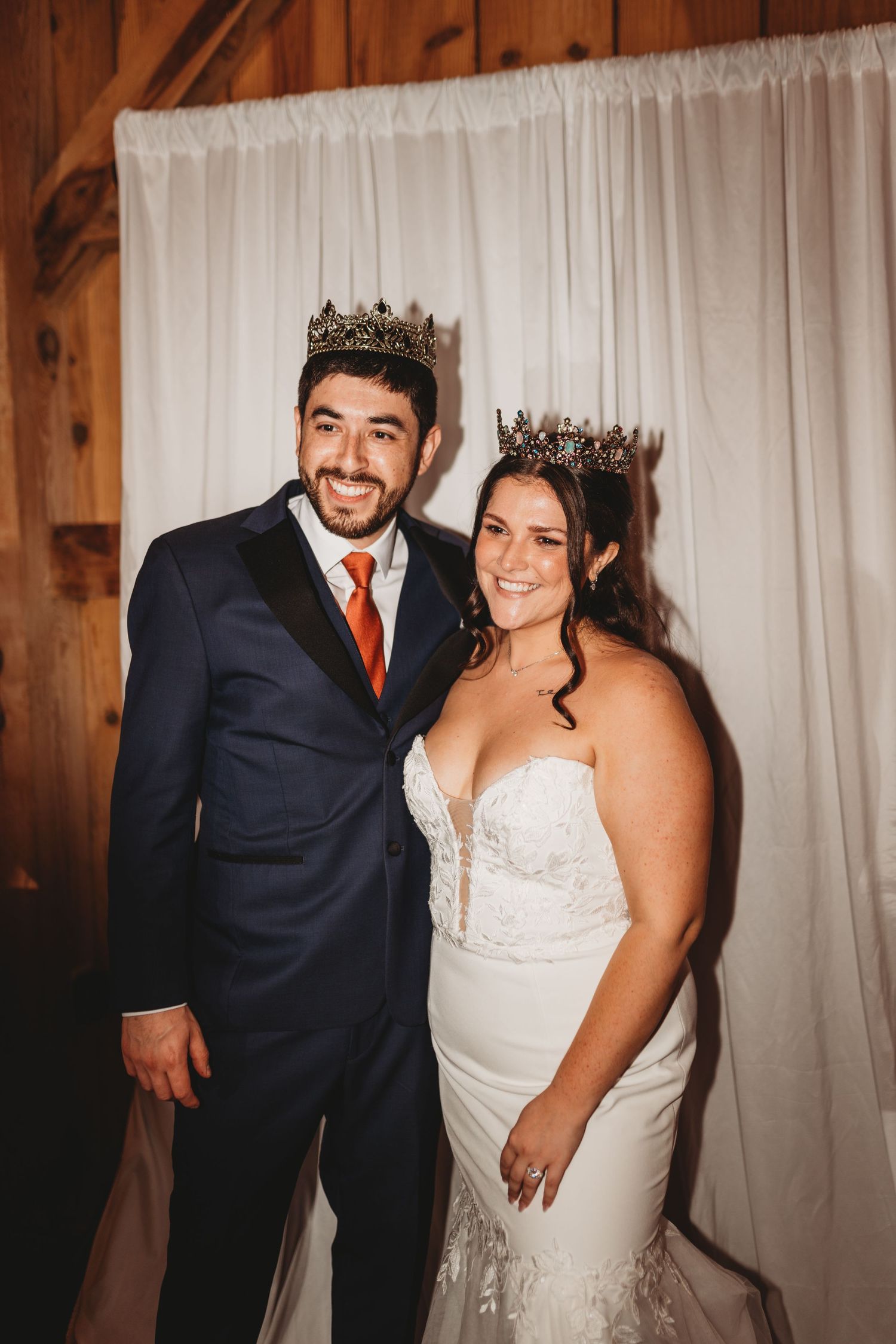 Regal couple wearing matching crowns pose against white backdrop in rustic barn setting.
