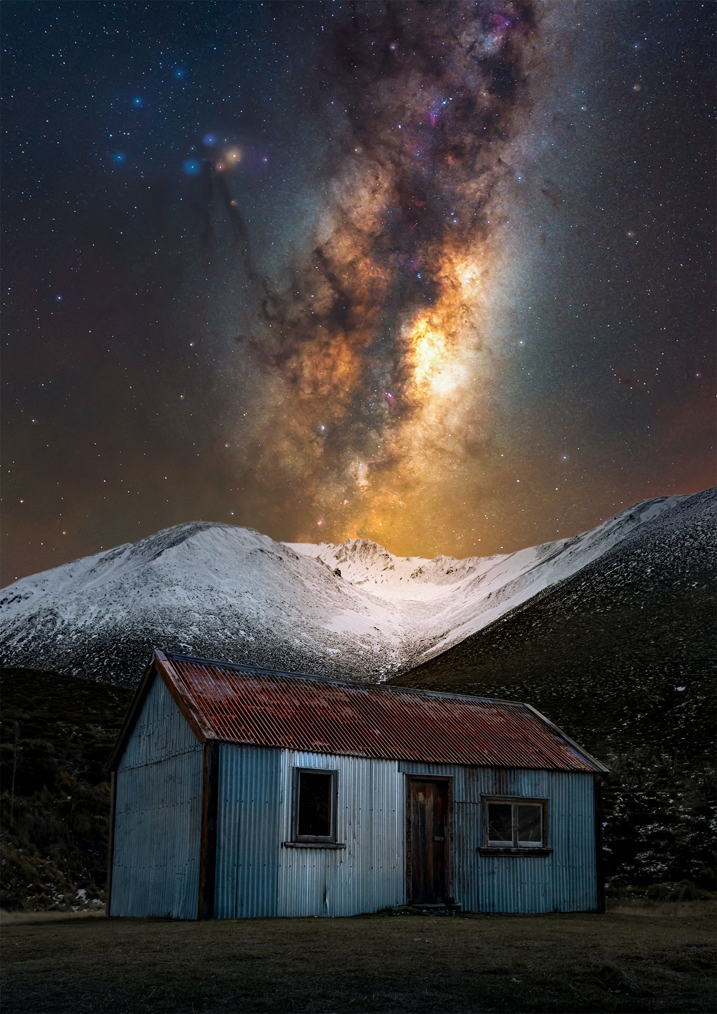 Old weathered cabin beneath a stunning starry night sky and snow-covered mountains.