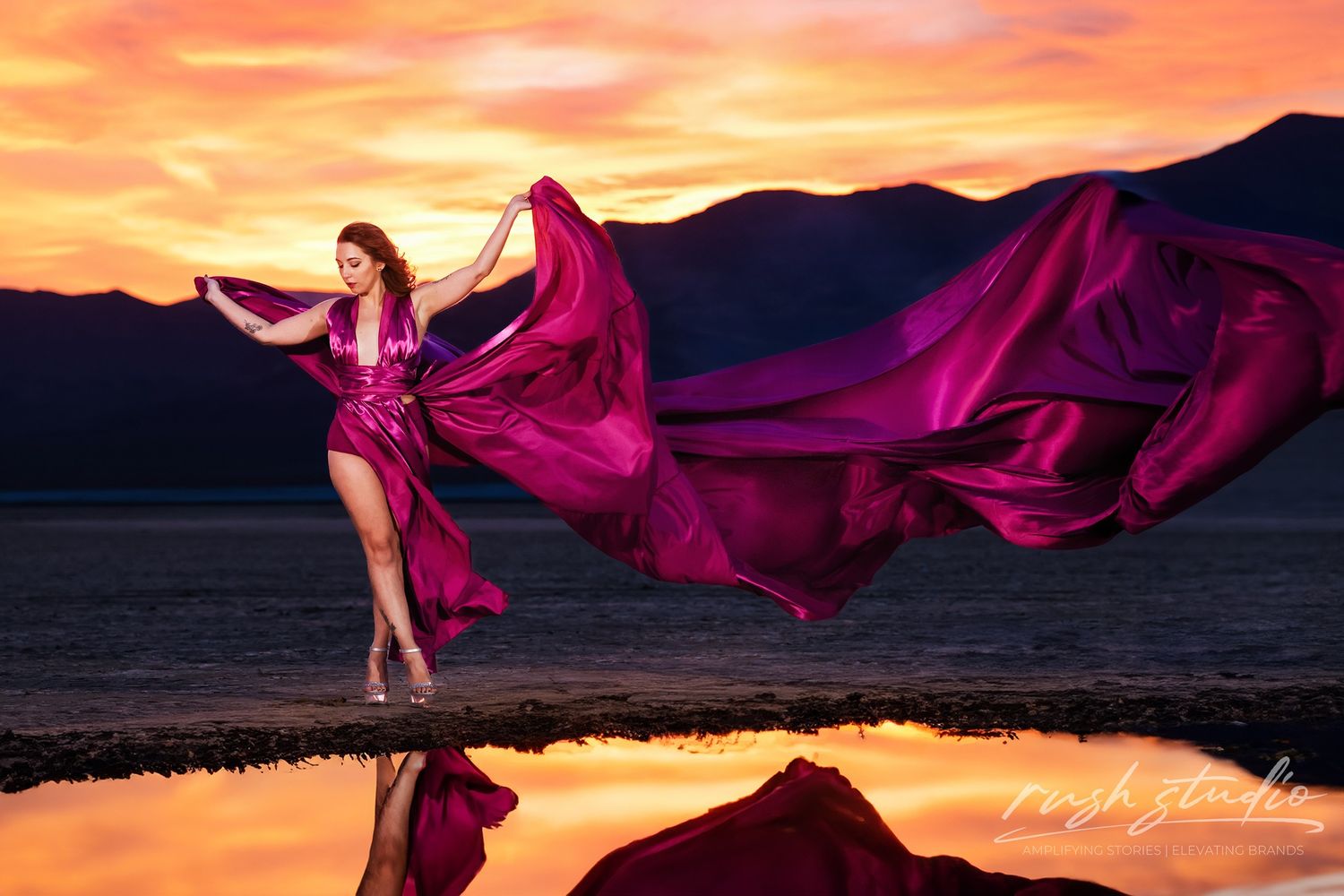 Flowing magenta dress billows dramatically against sunset mountains and reflective water.