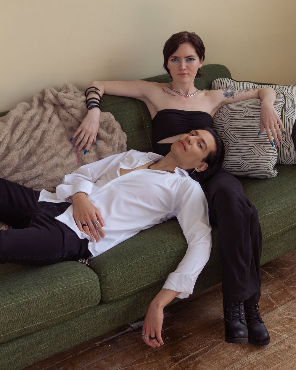 Two people lounging on a dark green velvet couch with decorative pillows and a fur throw in a cozy living room setting.