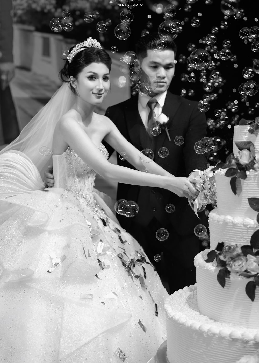 Black and white photo of couple cutting wedding cake surrounded by floating bubbles.