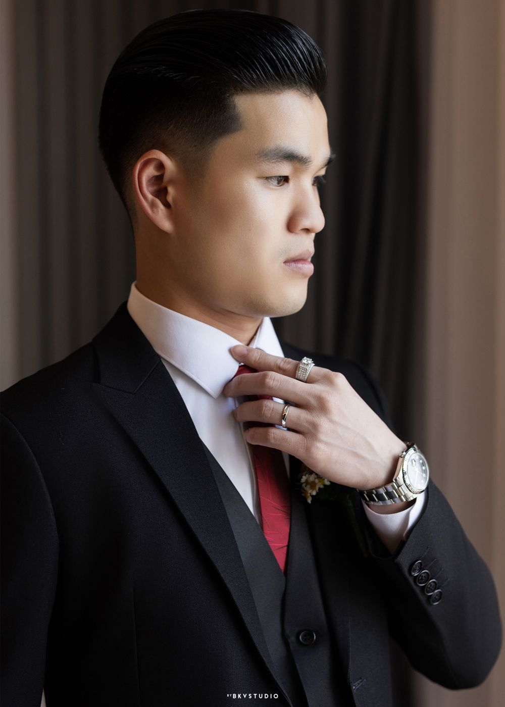 Formal portrait of groom in black suit adjusting red tie while wearing luxury watch.